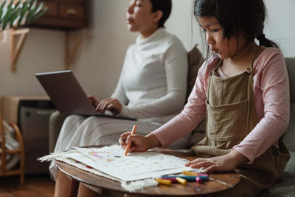 focused girl drawing on paper near mother with laptop